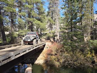 Image shows a jeep crossing a wooden bridge over a wooded creek.