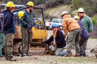 Image shows a group of men in hard hats kneeling around a piece of road construction equipment.