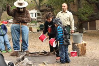 Image shows Smokey Bear advising children on how to put out a campfire safely while Forest Service staff look on.