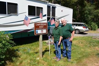 A man and woman campground host volunteer stand together beside an RV and a sign that says "Campground Host"