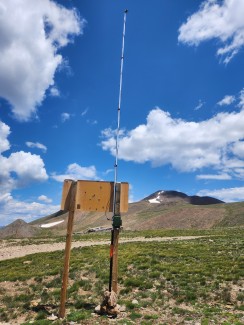 An antenae that is attached to the cack of a sign rises up into the blue sky above the mountain.