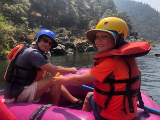 An adult and child on a whitewater raft on a river. Both are looking back to the camera