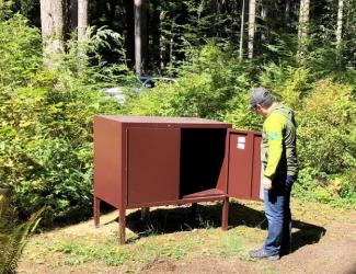A person stands in front of an open bear resistant metal container.