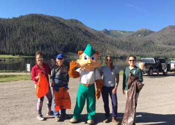 Woodsy Owl stands in a parking lot with two people on each side. A lake and mountains are in the background.
