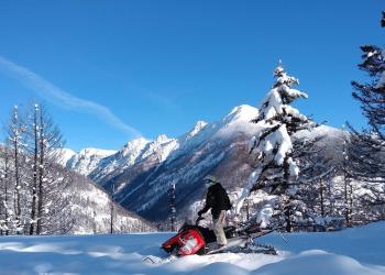 Man rides snowmobile with snow and tree covered mountains in the background.