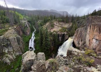 Two waterfalls drop from a a cliff area. The forest and mountains above blend in to a cloudy sky.