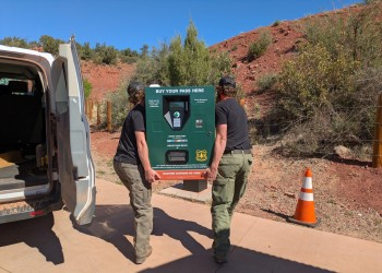 Two people wearing green pants and black t-shirts unload a large green machine from the back of a white van.