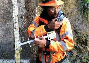 Man in orange jacket and hard hat using a measuring tool.