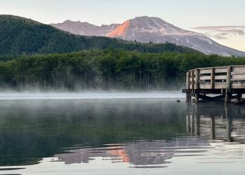This image shows aa mist covered lake and boardwalk trail with Mount St Helens in the background. Johnston Ridge itself is blocking a total view of the mountain.