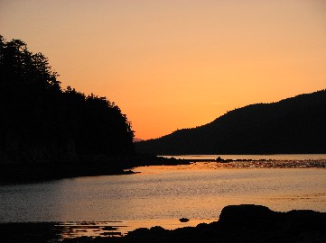 An orange sunset fills the sky over a bay amidst forested islands.