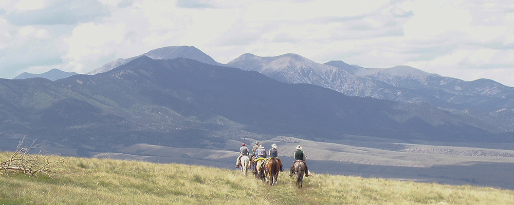 People riding horses over the crest of a mountain with larger mountains in the Background