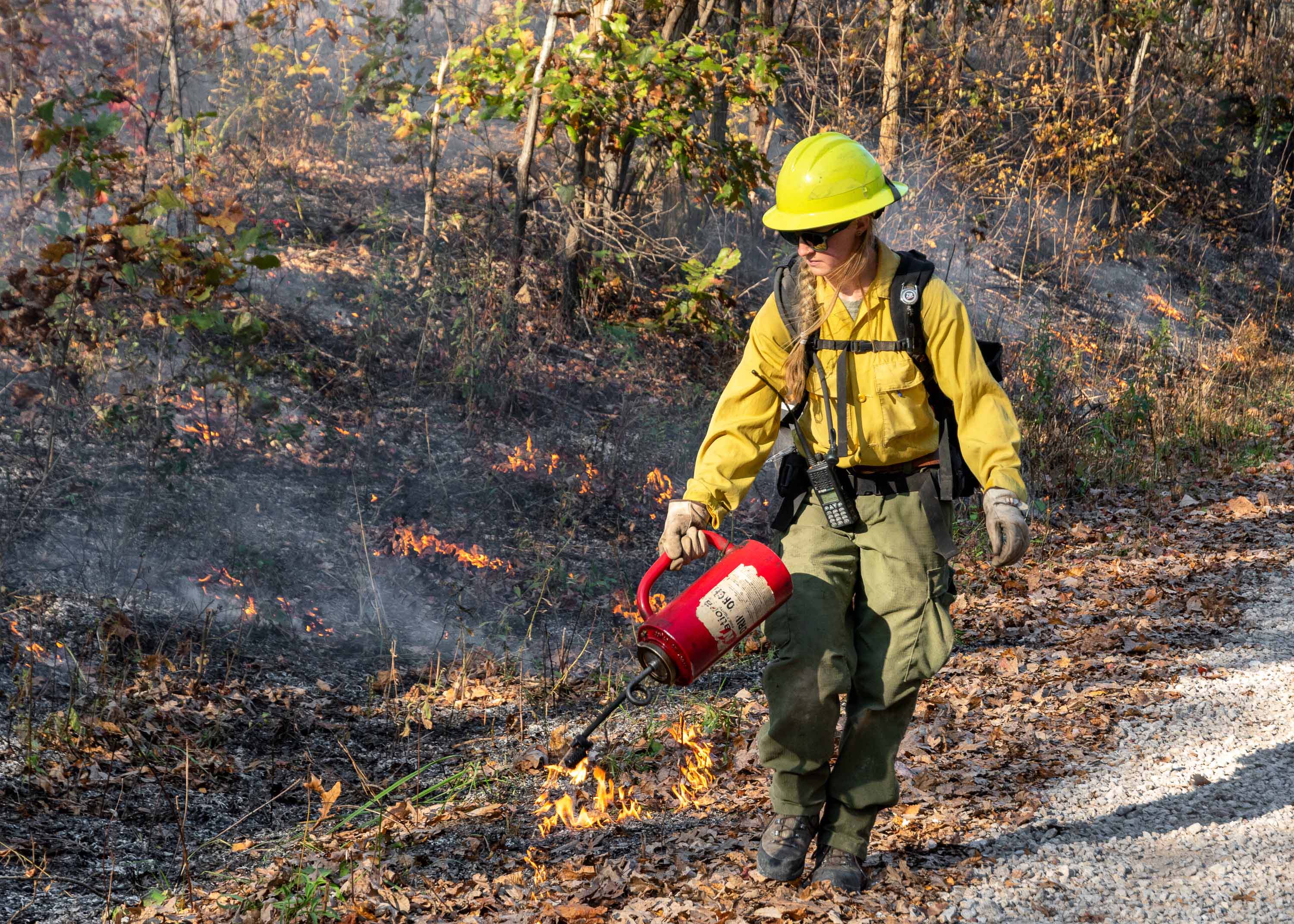 Employee in yellow and green firefighting gear uses a drip torch to conduct a prescribed burn.
