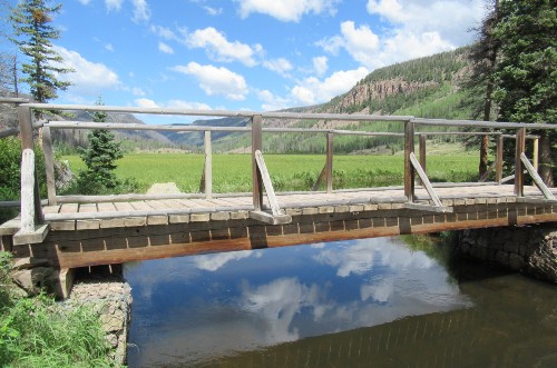 White clouds in a blue sky are reflected in a stream with a bridge through a green mountain valley.