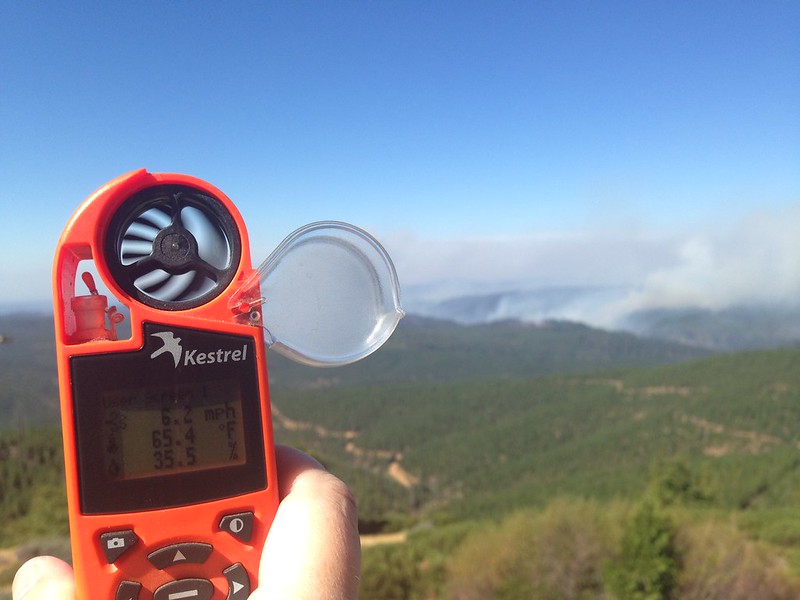 Hand holds up wind and weather meter to gauge fire conditions.