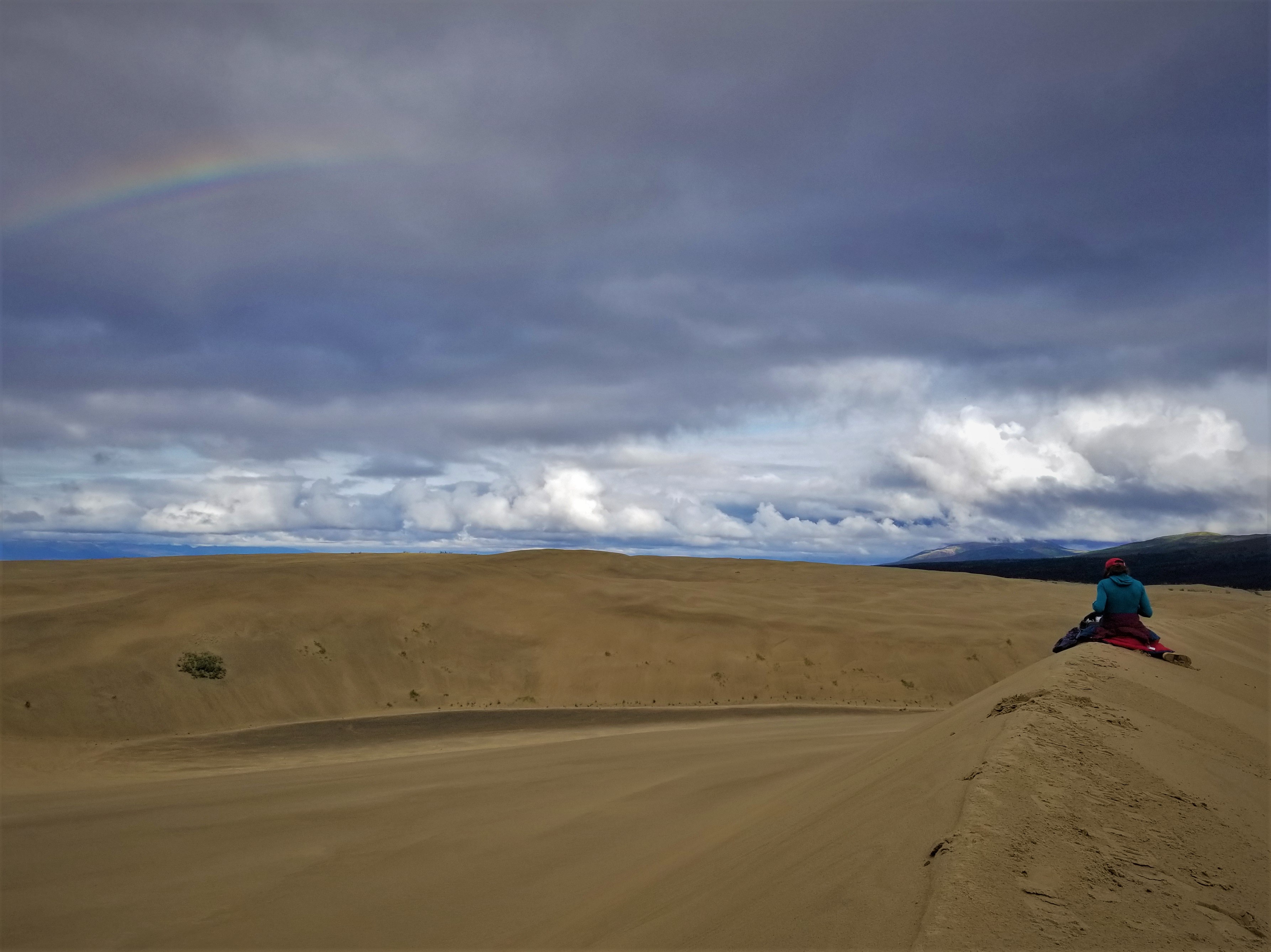 Artist Amy Martin sits on a sand dune under a rainbow in Noatak National Wildlife Refuge.