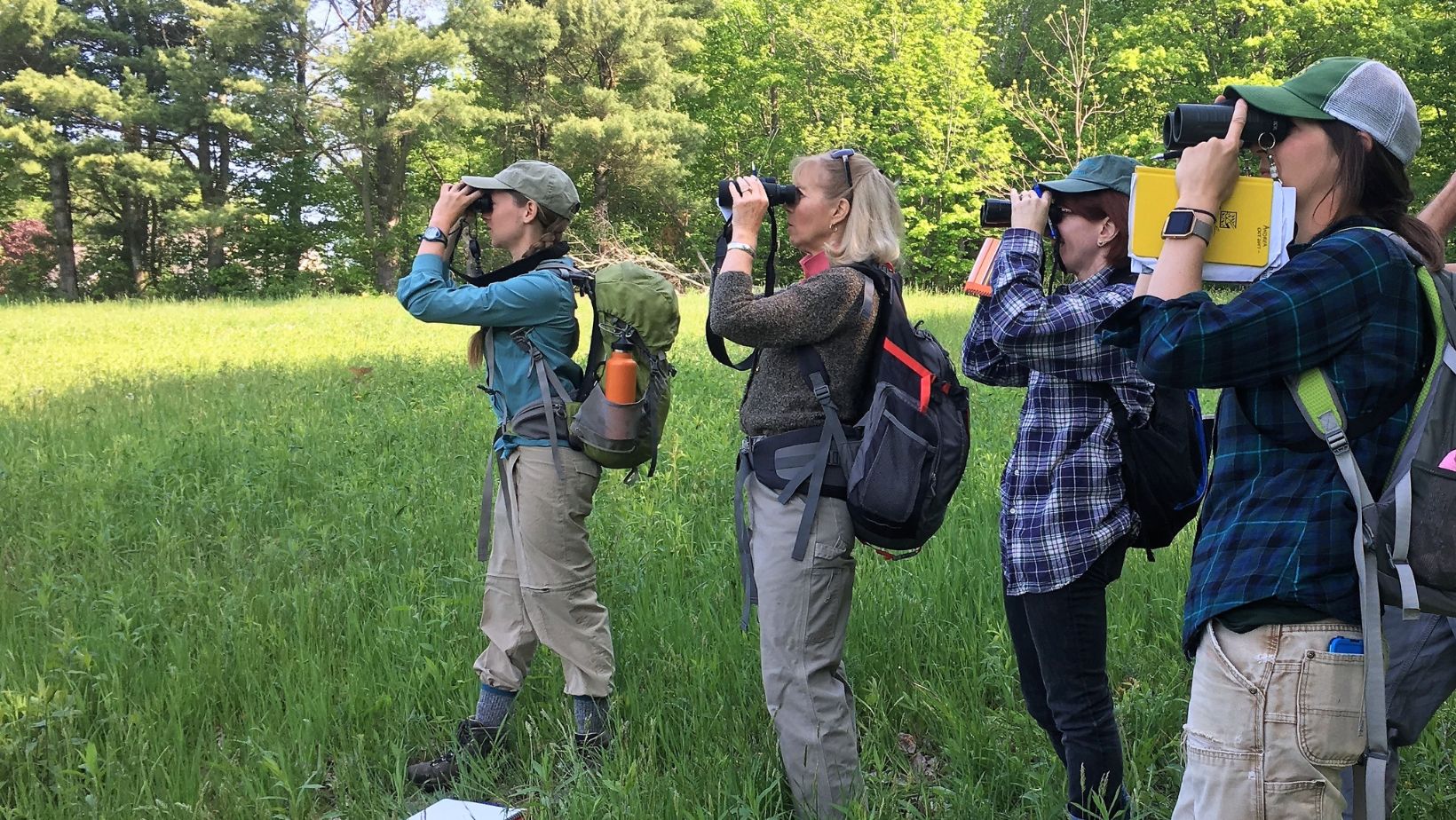 A group of women in outdoor gear looking towards the woods with binoculars