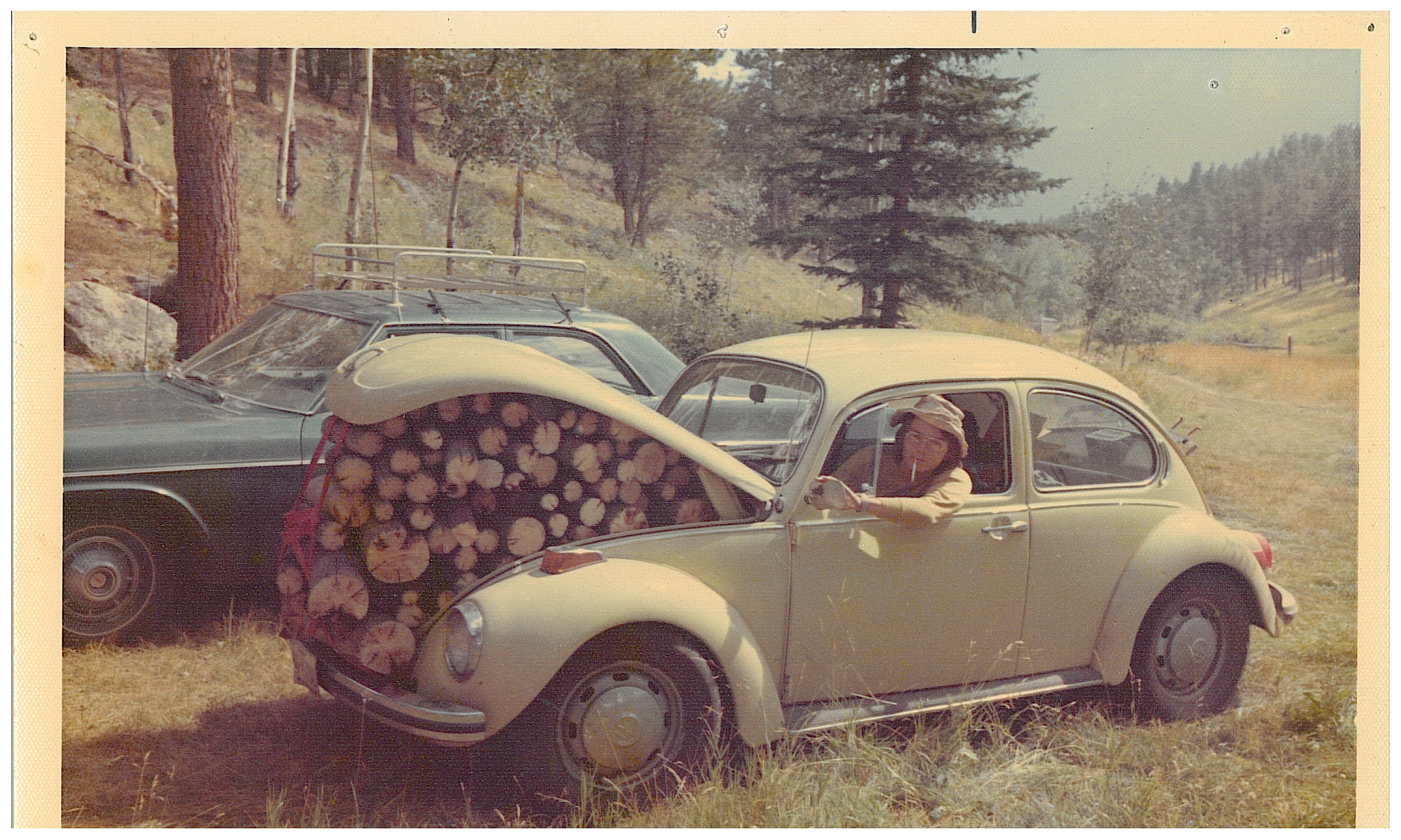 Historic photo of man smoking a cigarette leaning out a VW beetle window. Car is packed with firewood.