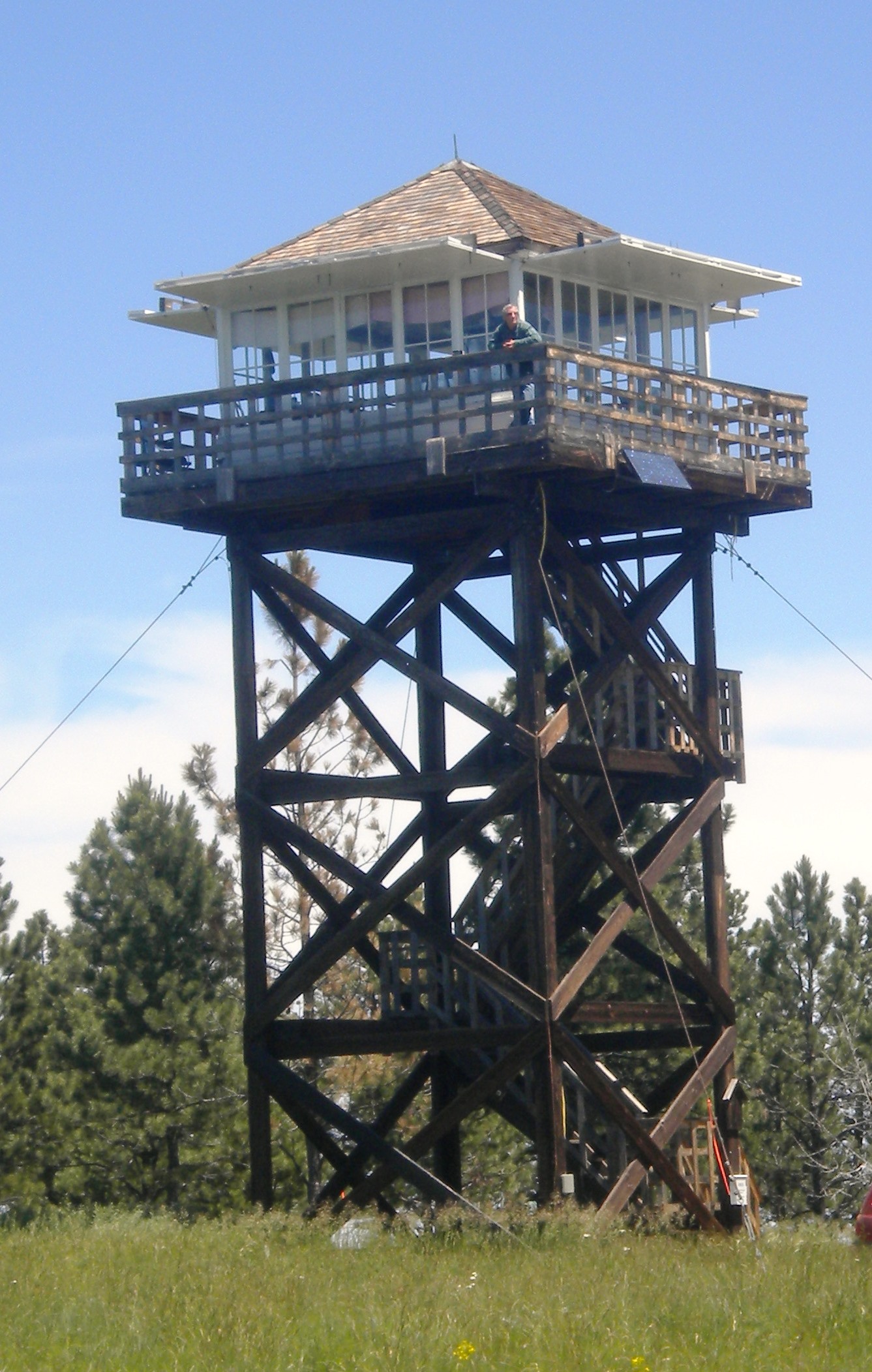 A photo of a fire lookout tower. A person is standing on the deck looking out from the tower. Pine trees are behind the tower.