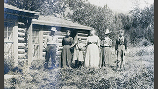 A black and white photo of a family of six people standing in front of a wood cabin.