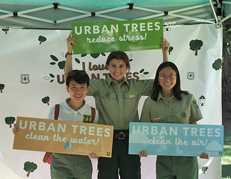 Three people hold signs saying “Urban trees... reduce stress, clean the water, and clean the air.