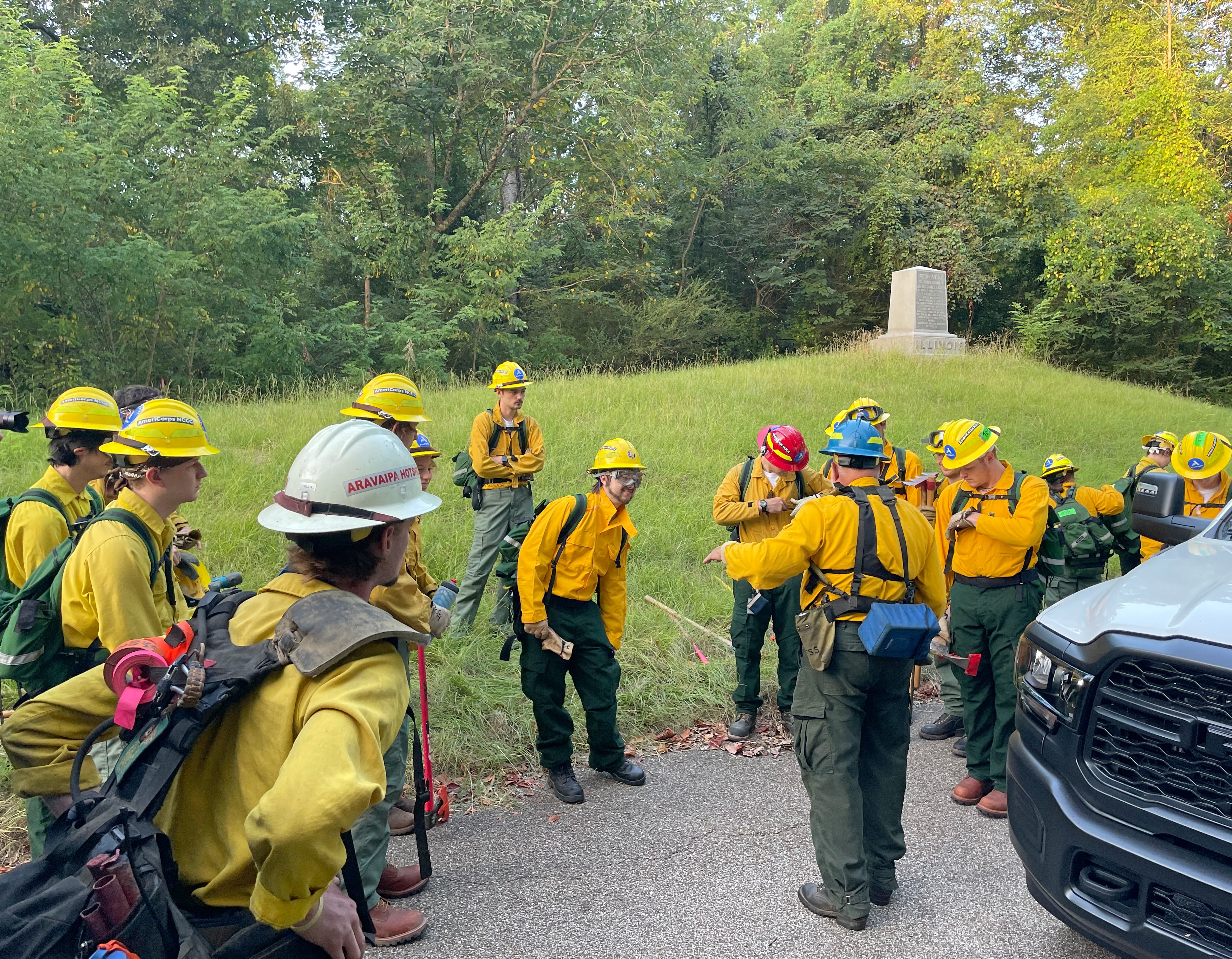 A group of young adults wearing firefighter gear gather outside in a forest listening to a man talk.