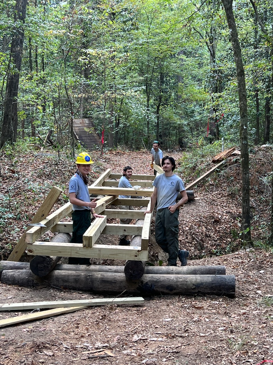 Four young adults wearing AmeriCorps t-shirts building a trail foot bridge in a forest.
