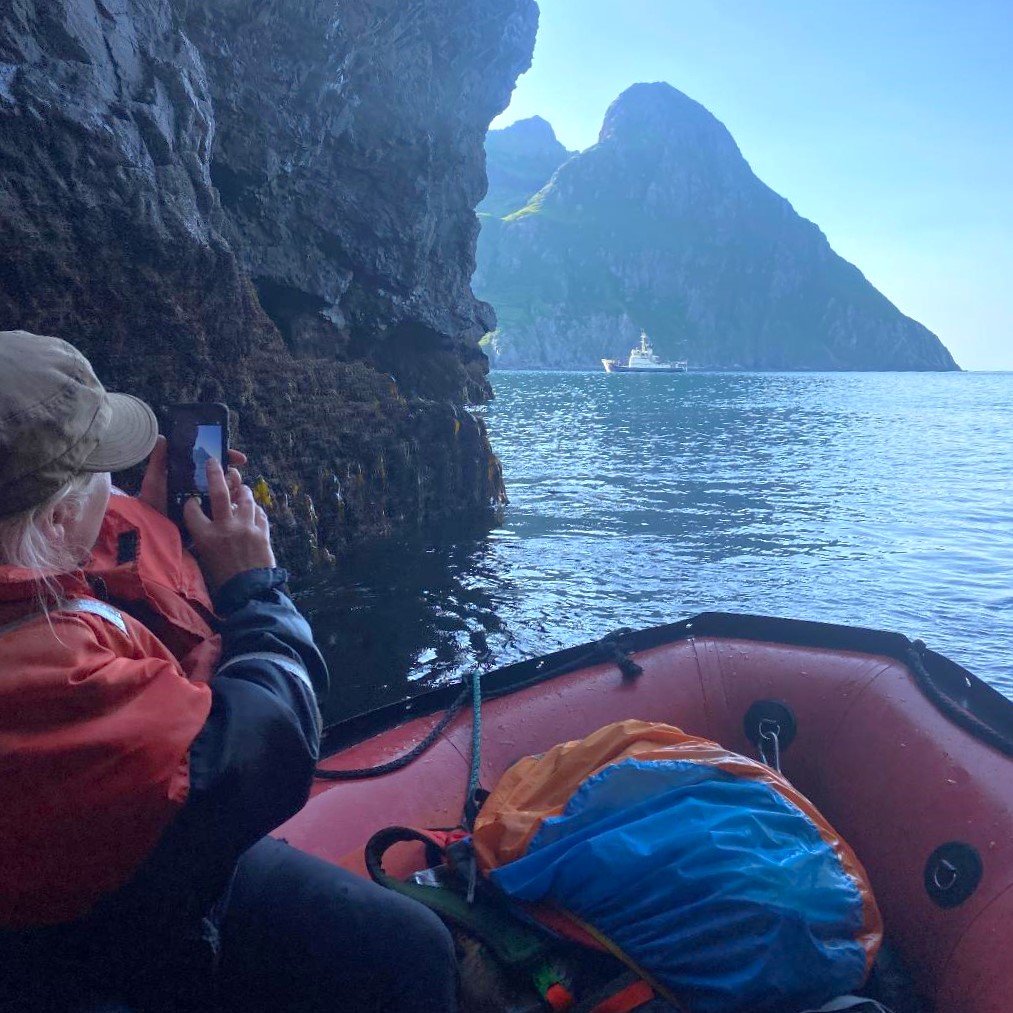 Bridget Bruner, sitting in a pack raft, points a cell phone camera at the RV Tiglax in the Aleutians