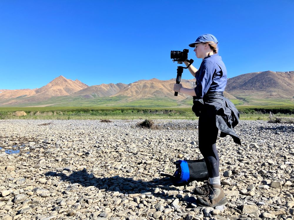 Rachel Heckerman looks through a camera, standing on a rocky landscape surrounded by mountains.