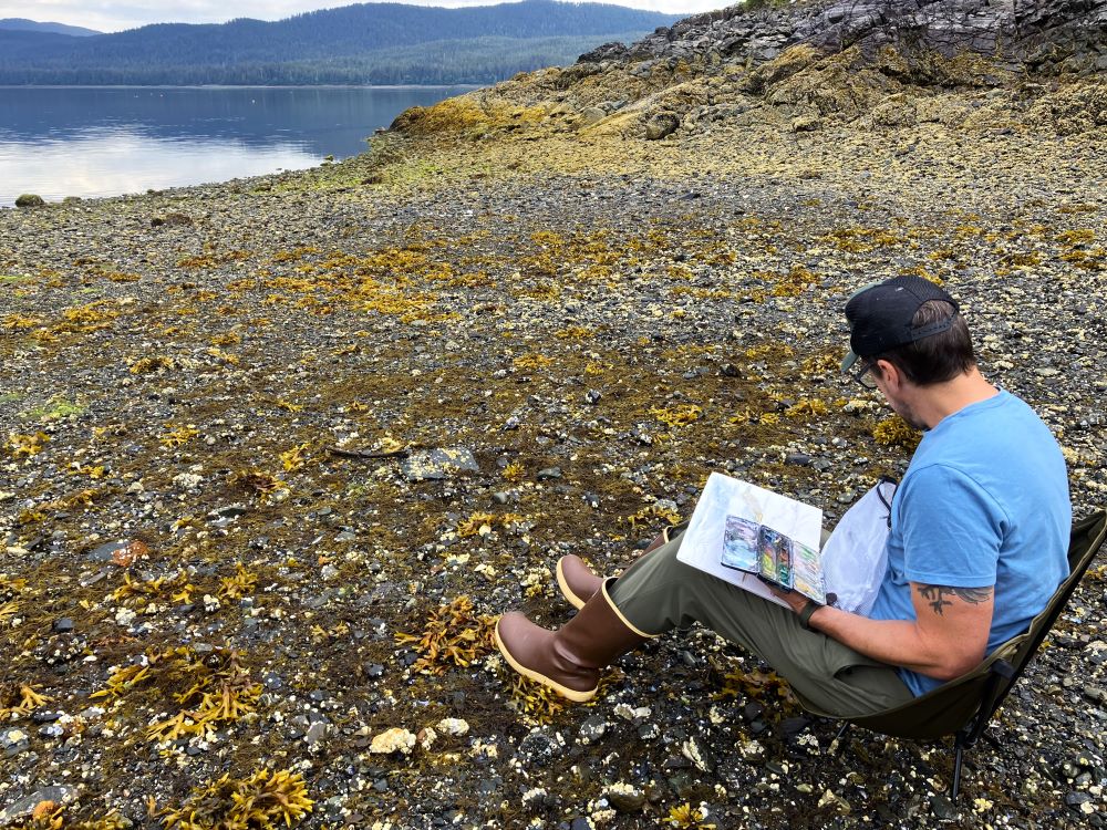 Tom Hanicak sits in a camp chair on a rocky beach painting with water colors.