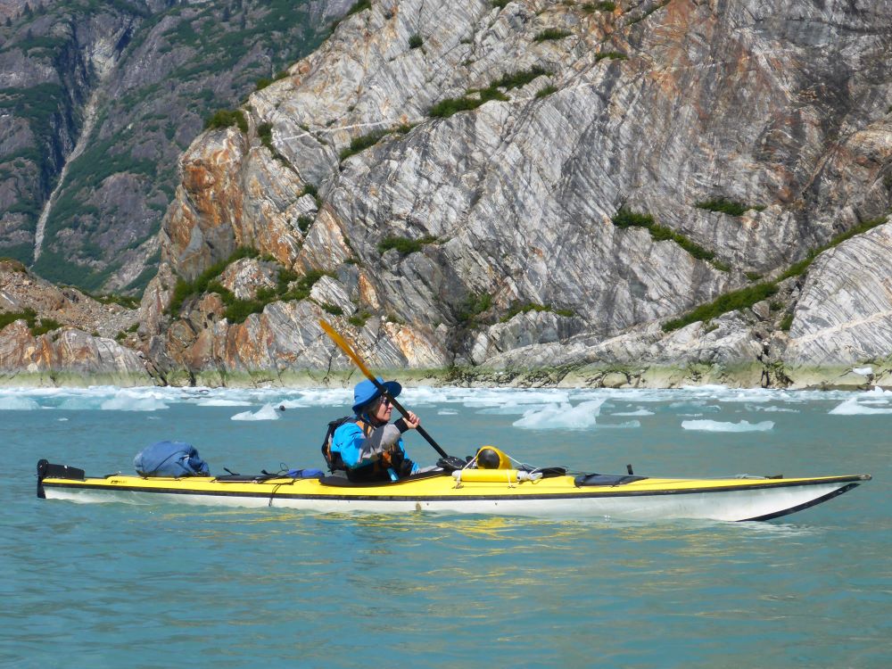 Youpa Stein paddles a sea kayak past steep granite cliffs and icebergs of a fjord.