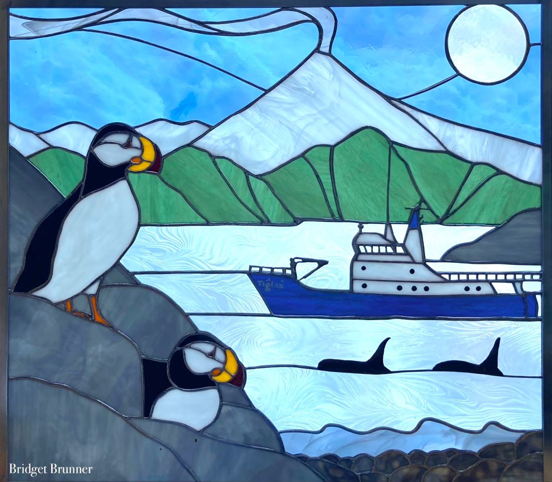 Stained glass work with two horned puffins on rocks, the research vessel Tiglax, two orca, mountains