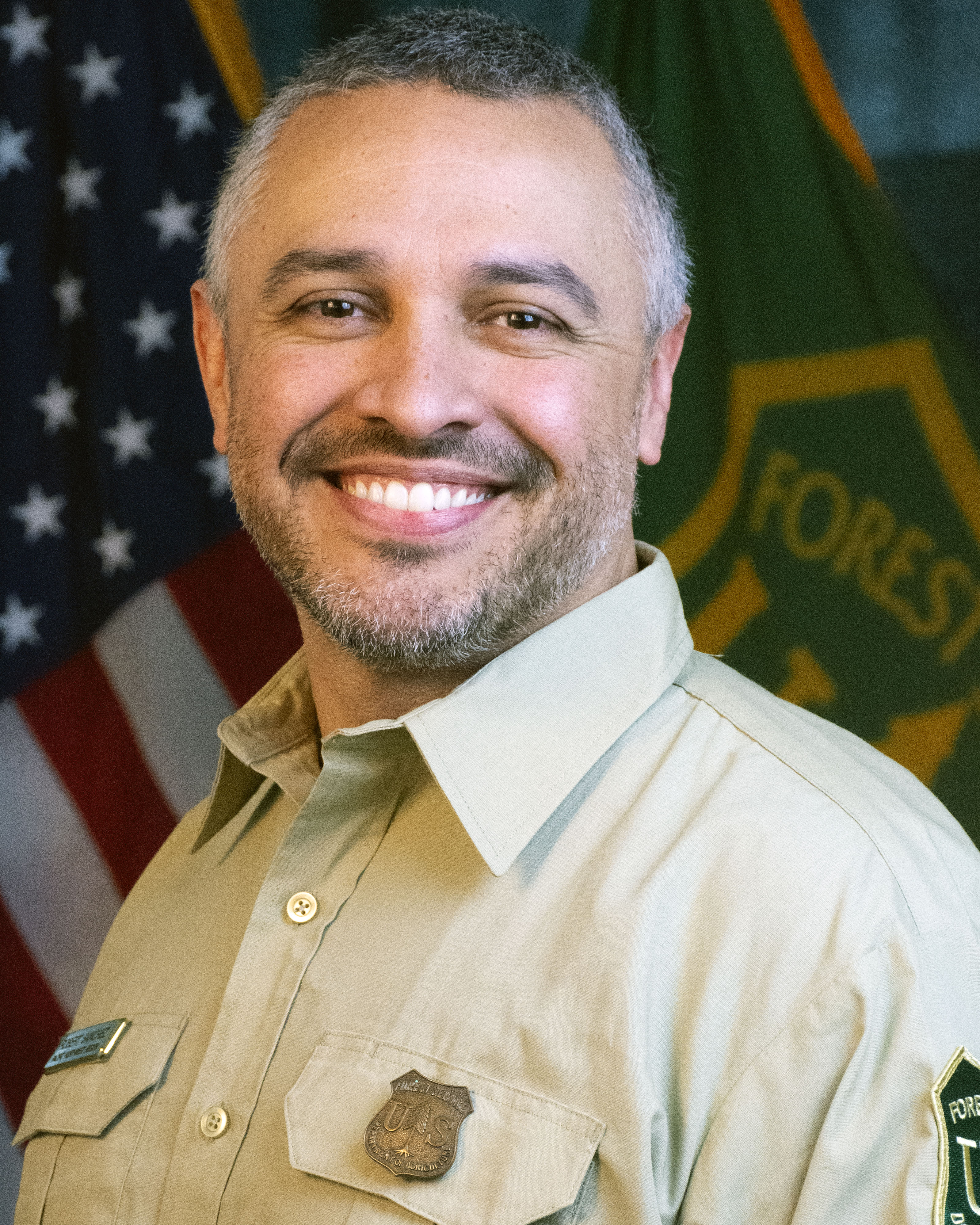 portrait of a man in a forest service uniform with the US and Forest Service flags behind him