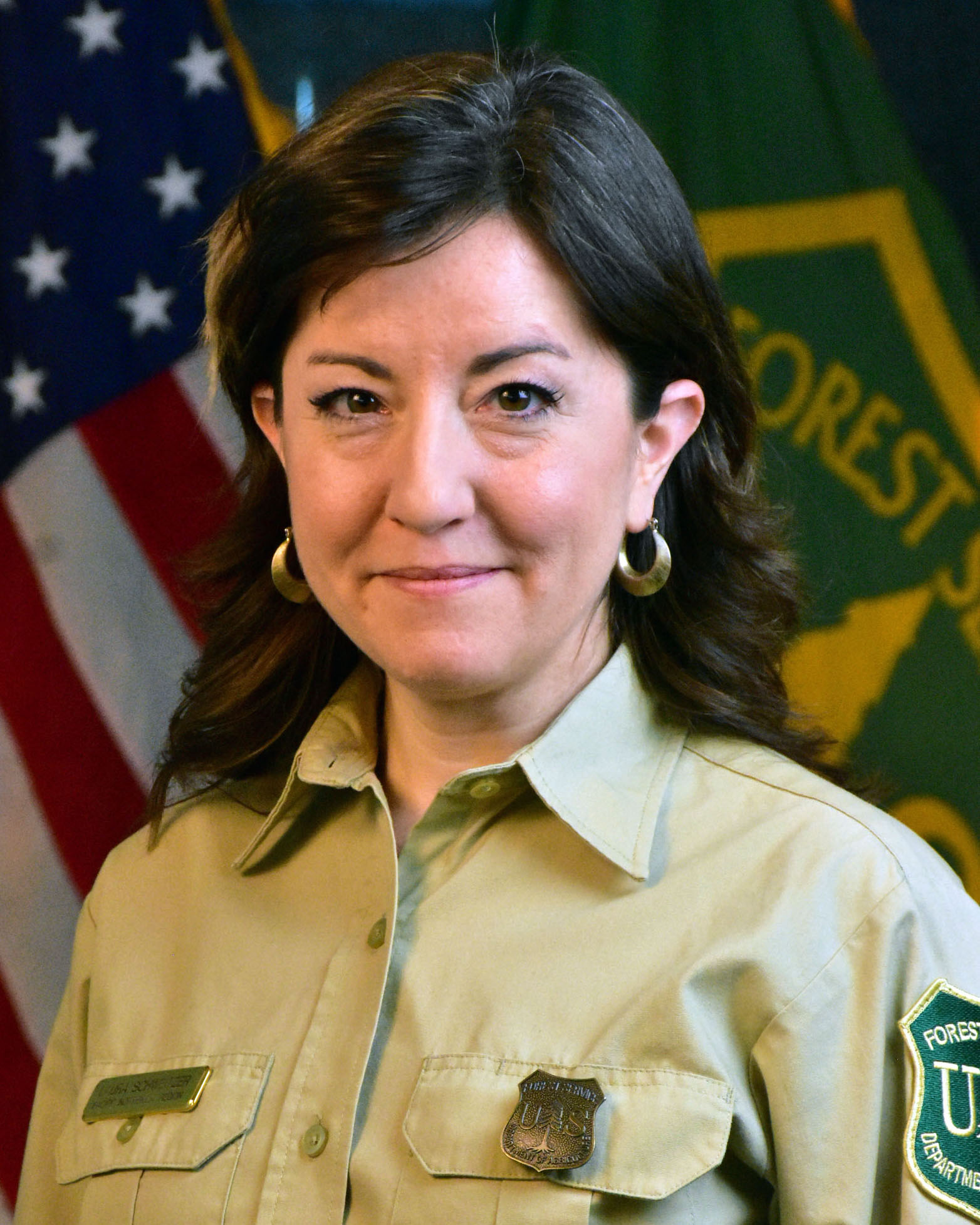 A portrait of a women in the forest service uniform with US and forest service flag behind her.