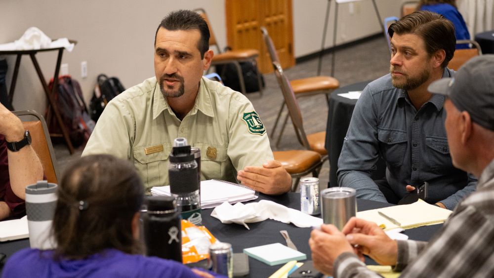 A Forest Service employee in uniform talks to people sitting at a table.