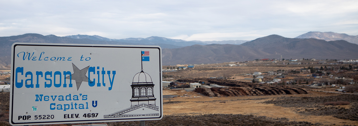 Sign saying “Welcome to Carson City, Nevada’s Capital” with brown earth and mountains behind.