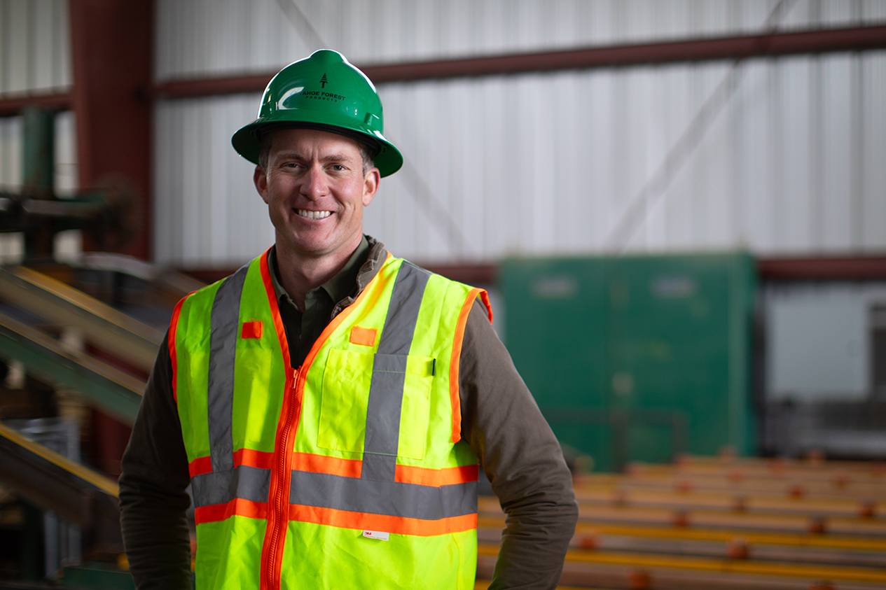 Man wearing green hard hat and safety vest with metal building and lumber in background.