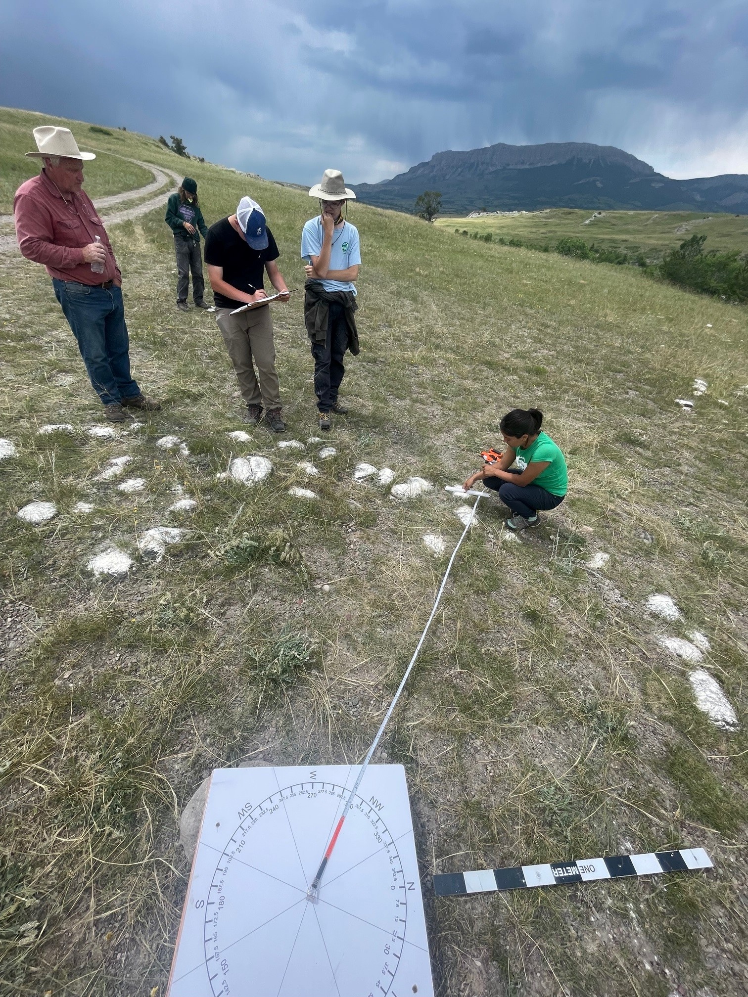 A photo of a group of people outdoors. One person is sitting on the ground, a measuring tape and other tools.