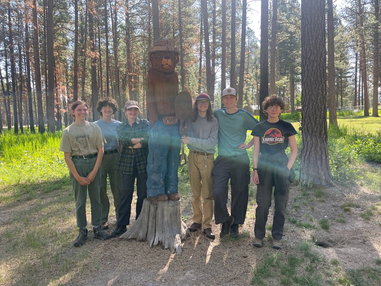 Six people standing outdoors next to a smokey bear carving. Trees in the background.