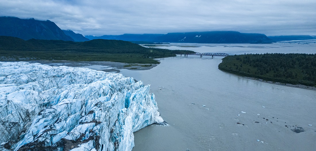 The toe of a massive glacier extends into the Copper River Delta, a bridge is in the distance.
