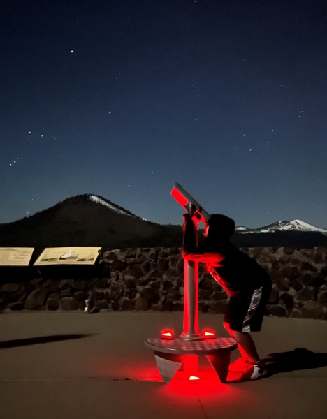 A young boy looks through a scope at night with mountains and stars in the distance.