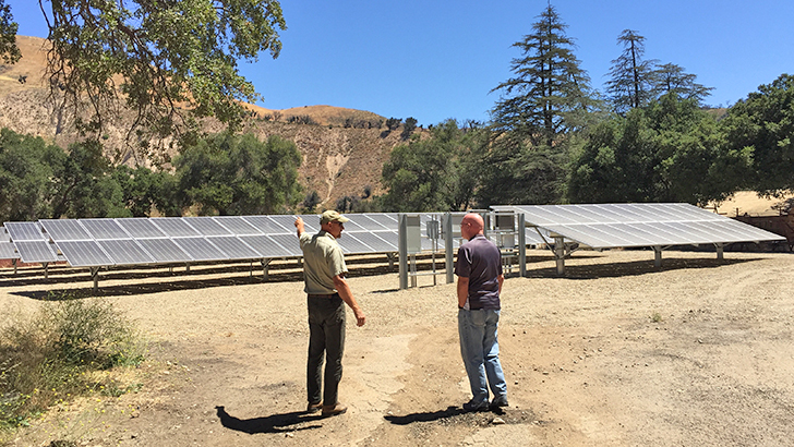 Two men stand in a dirt field filled with solar panels.