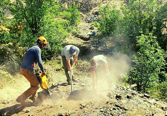 Three workers dig into a dusty trail with tools.