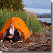 Jennifer sitting next to her tent near the shore in the Tebenkof Bay Wilderness.