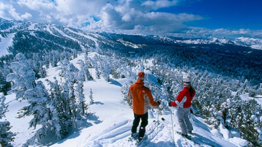 Two skiers stand on a snow covered peak in downhill ski gear.
