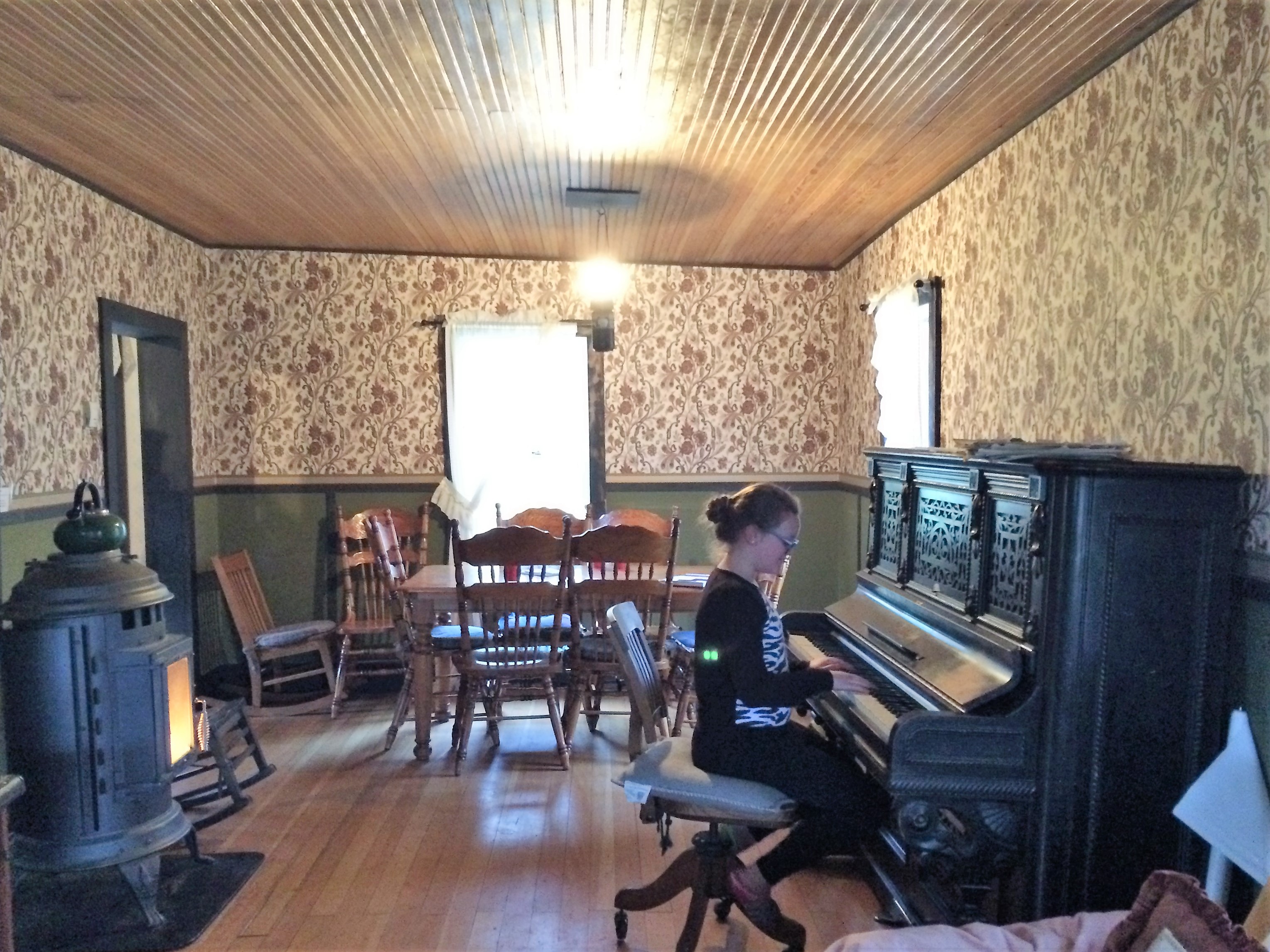A rustic living room and dining room with wood floors, an old-fashioned warming stove, piano, and opulent wallpaper.