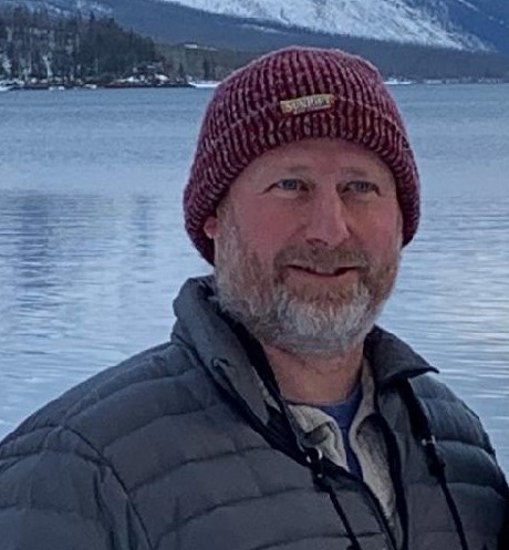 Close up of a smiling man with a short gray beard in a beanie and down coat in front of a lake.