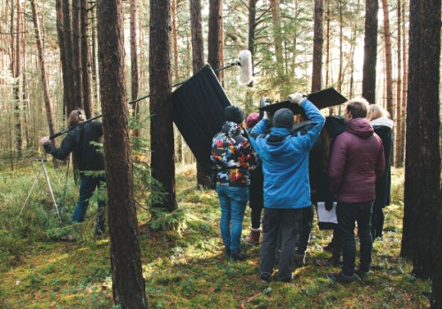 A filming crew with microphones and cameras films in the forest under the trees.