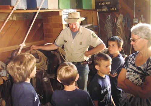 A mule packer with the Forest Service speaks to a small group of children in a stall or barn area.
