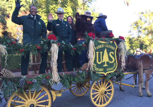 Forest Service wagon and mules participate in the Rose Parade in Pasadena with riders waving.