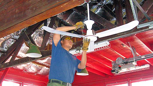 A man on a ladder uses tools to attach fan to ceiling.